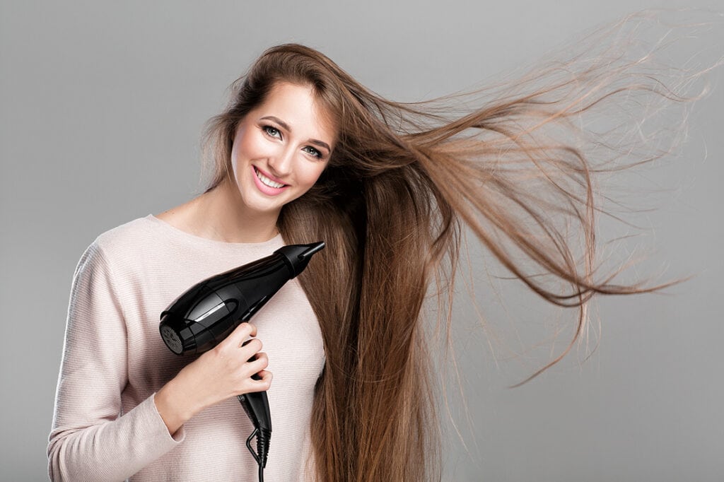 beautiful smiling woman drying her long hair with dryer over gray background